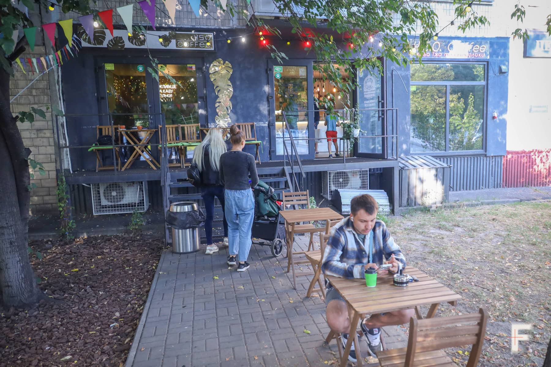 На передньому плані чоловік сидить за столом, п’є каву, курить цигарку та грає на телефоні; на задньому плані кав’ярня, жінка піднімається сходами з дитячим візком