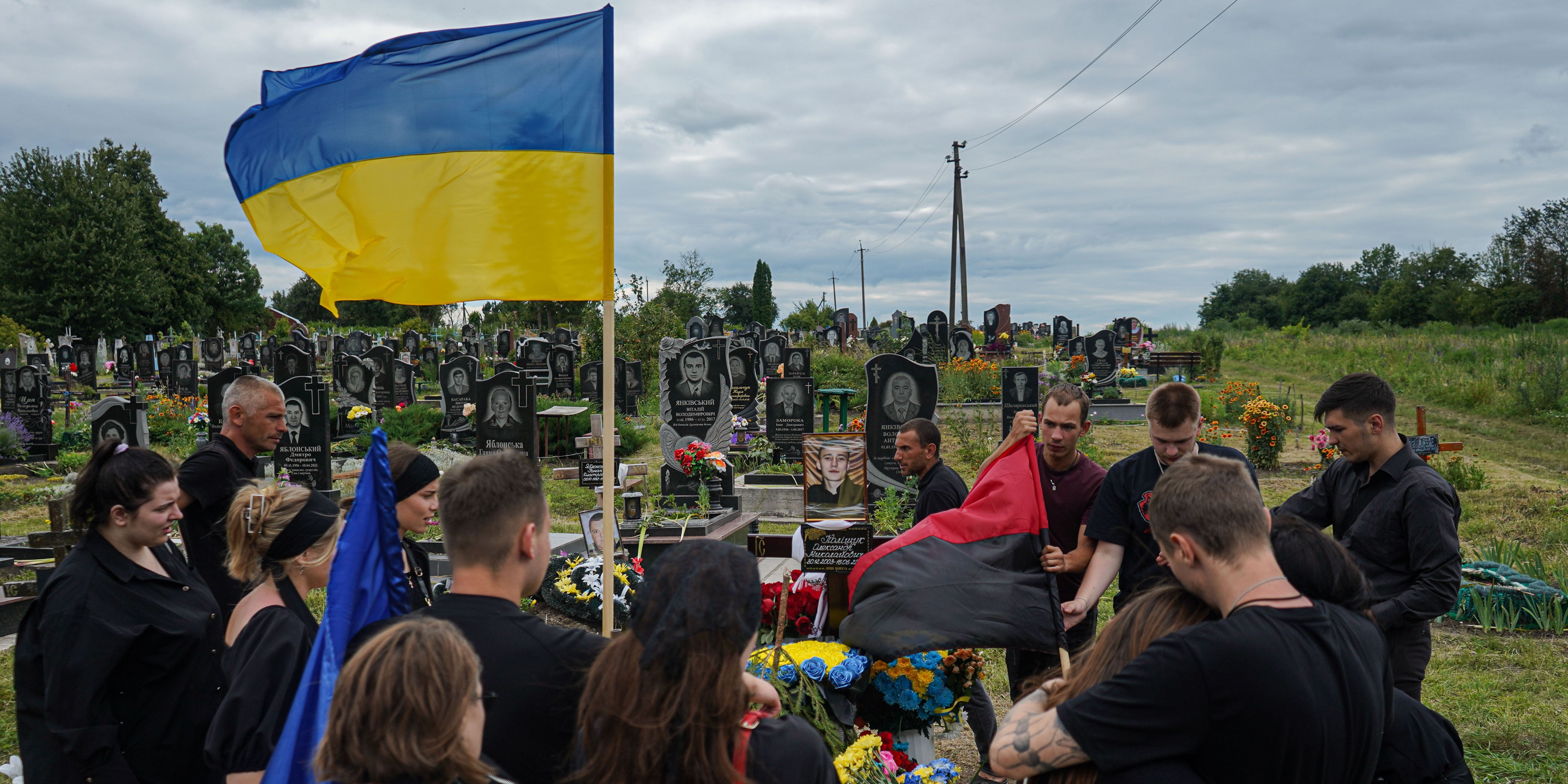 Група людей стоїть на кладовищі під час похорону, видно український прапор.