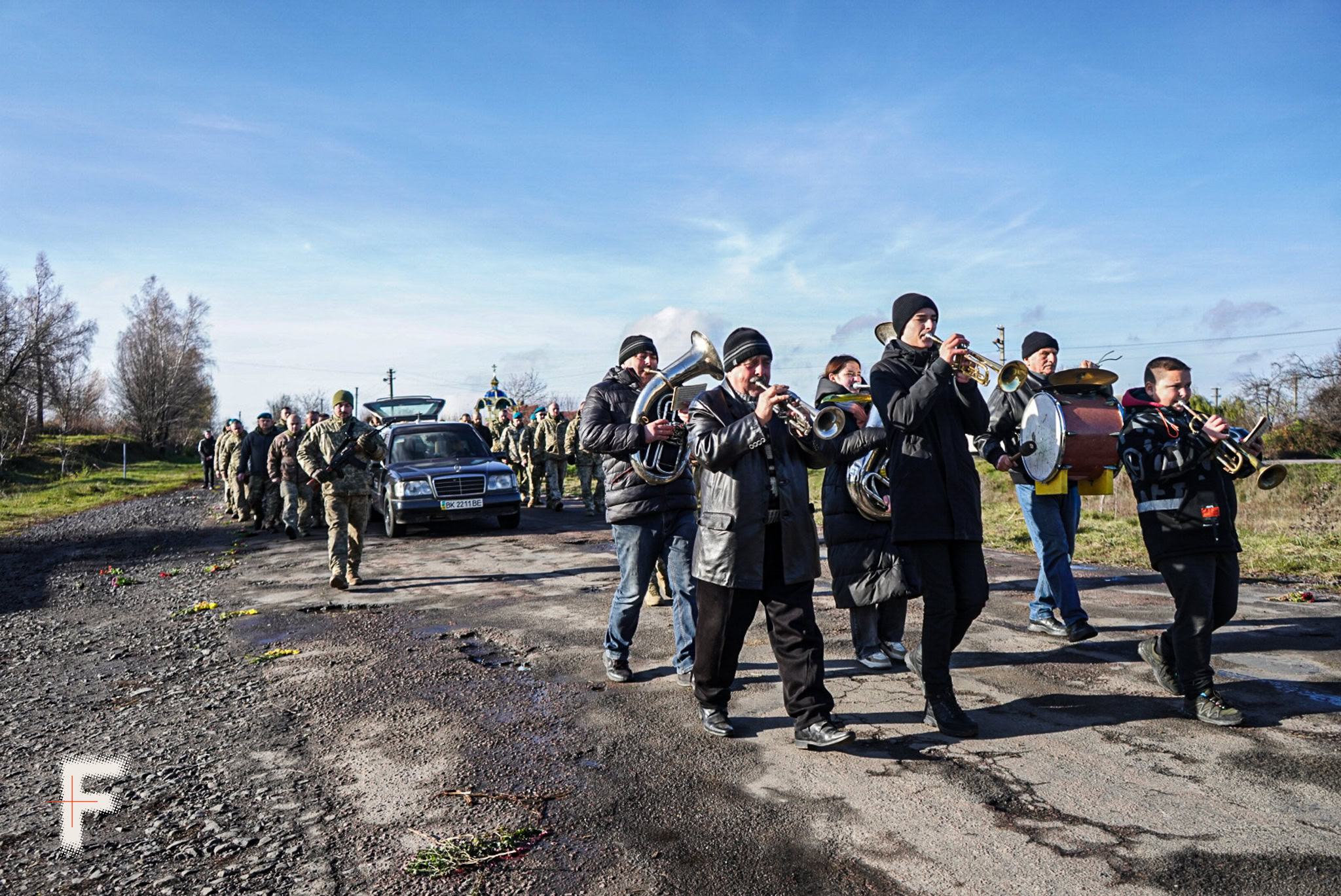 Похоронний оркестр, за яким їде машина з труною, поруч ідуть військові.