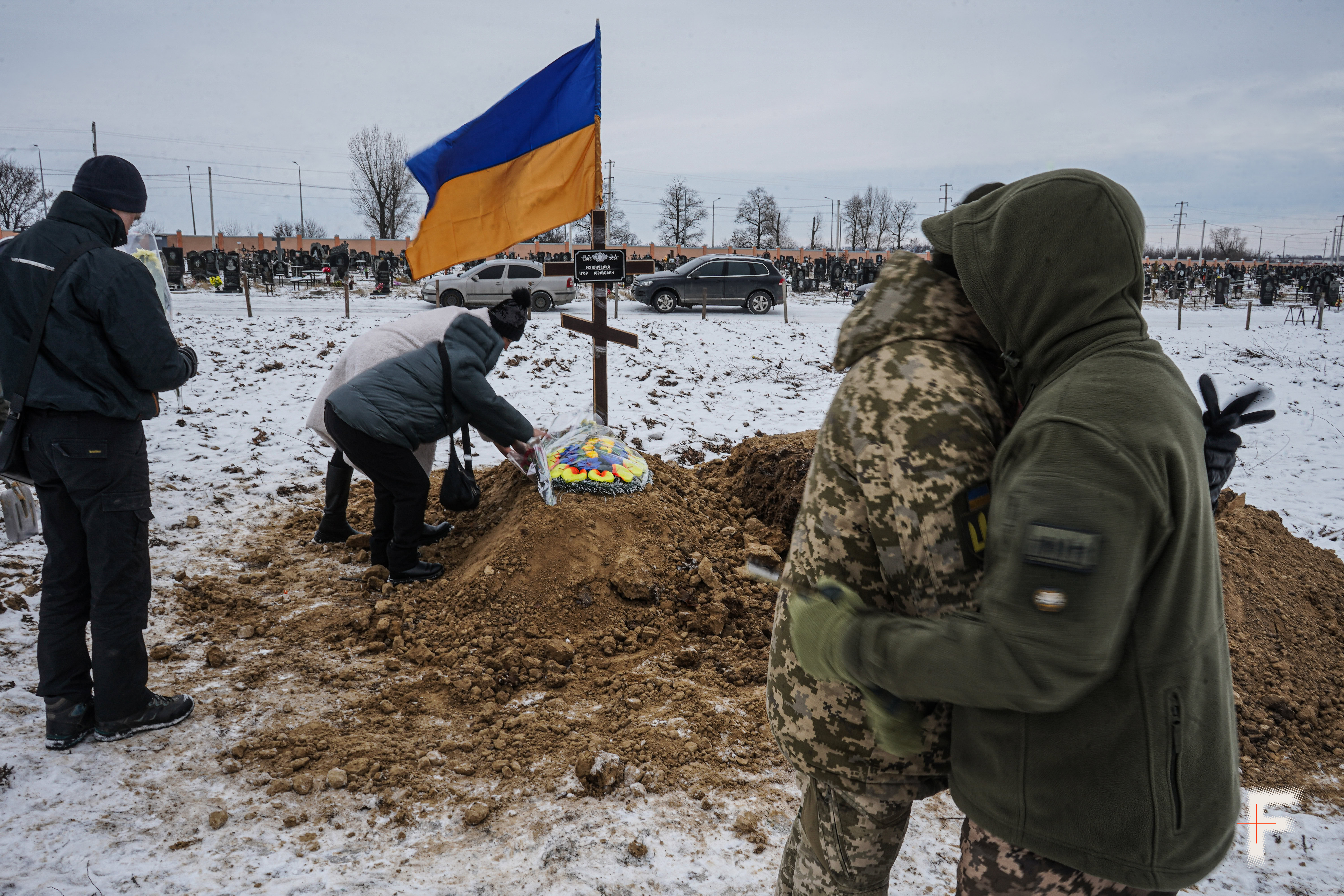 На передньому плані люди обіймають військового, на задньому плані видно могилу та сніг.