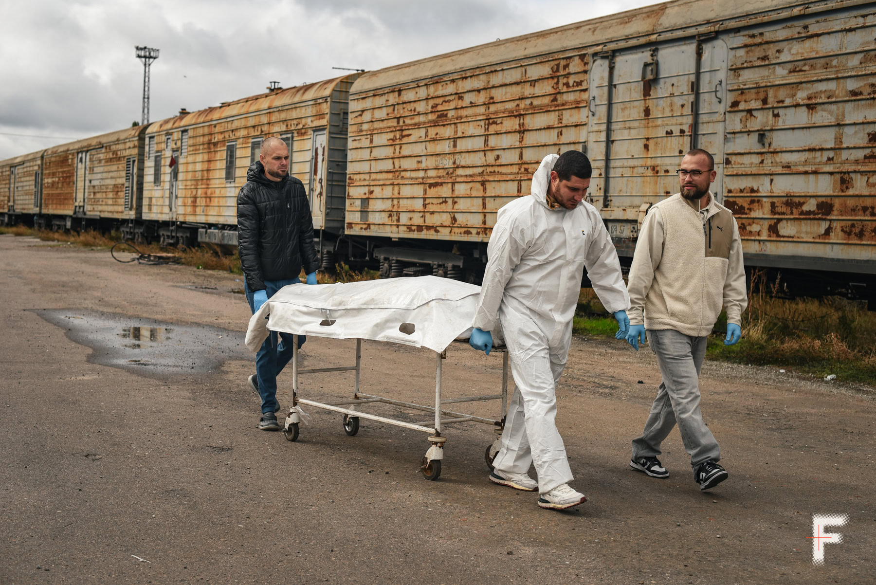 Троє чоловіків несуть білий пакет із тілом на тлі старого залізничного вагона.