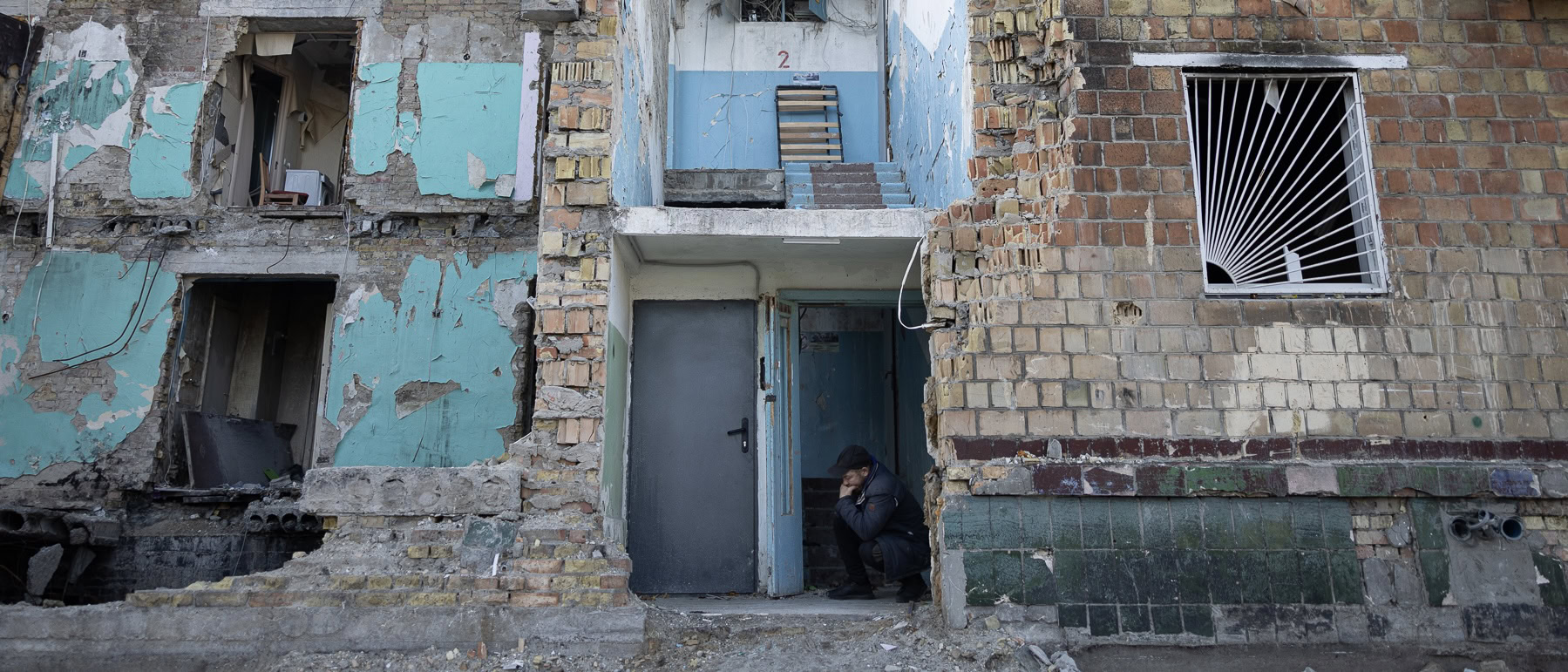 Чоловік присів у дверному проході зруйнованого будинку після обстрілу.
