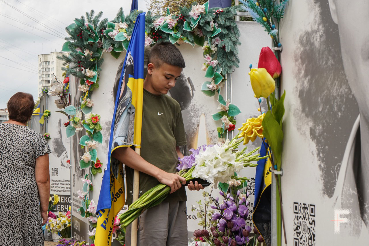 Молодий чоловік біля меморіальної стіни з портретами загиблих військових тримає прапор України та квіти, схиливши голову