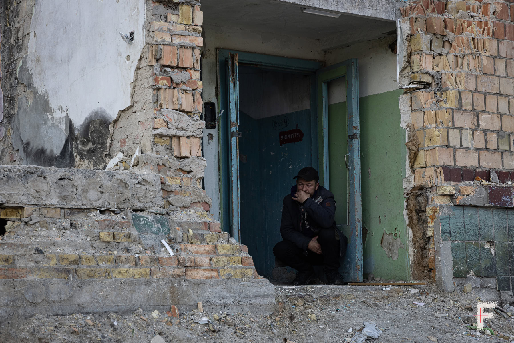 Чоловік, присівши, курить перед входом до будинку, зруйнованого бомбардуванням.