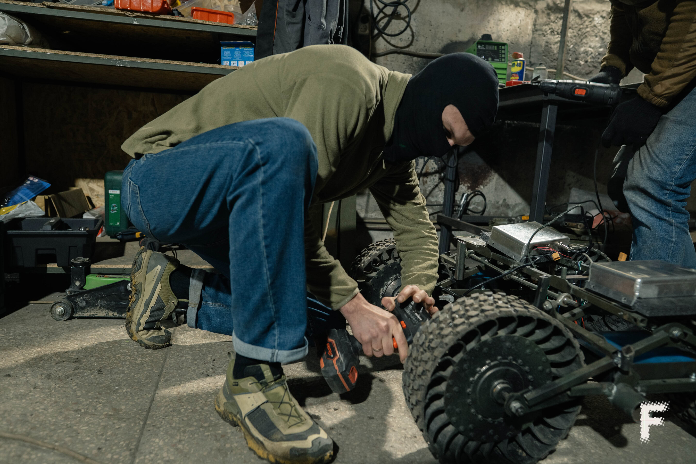 Військовий у балаклаві ремонтує наземного робота, закручуючи деталі корпусу в ангарі.