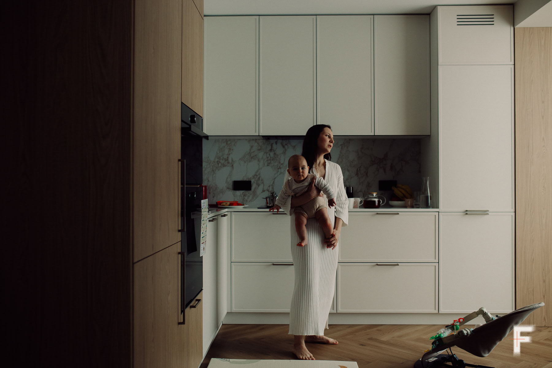 Жінка стоїть на кухні та тримає немовля на руках.