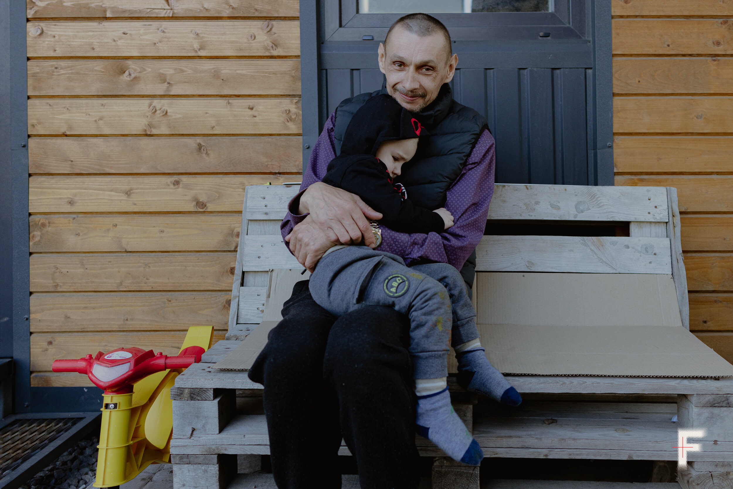 Чоловік сидить на лавці з піддонів і тримає маленького сина на руках, сімейне життя під час війни, Україна, 2026